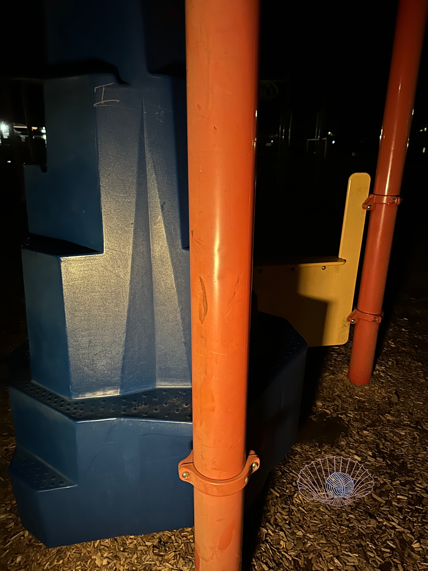 A section of playground set at night with red poles, a yellow partition, and a blue climbing piece. Wood chips are on the ground.