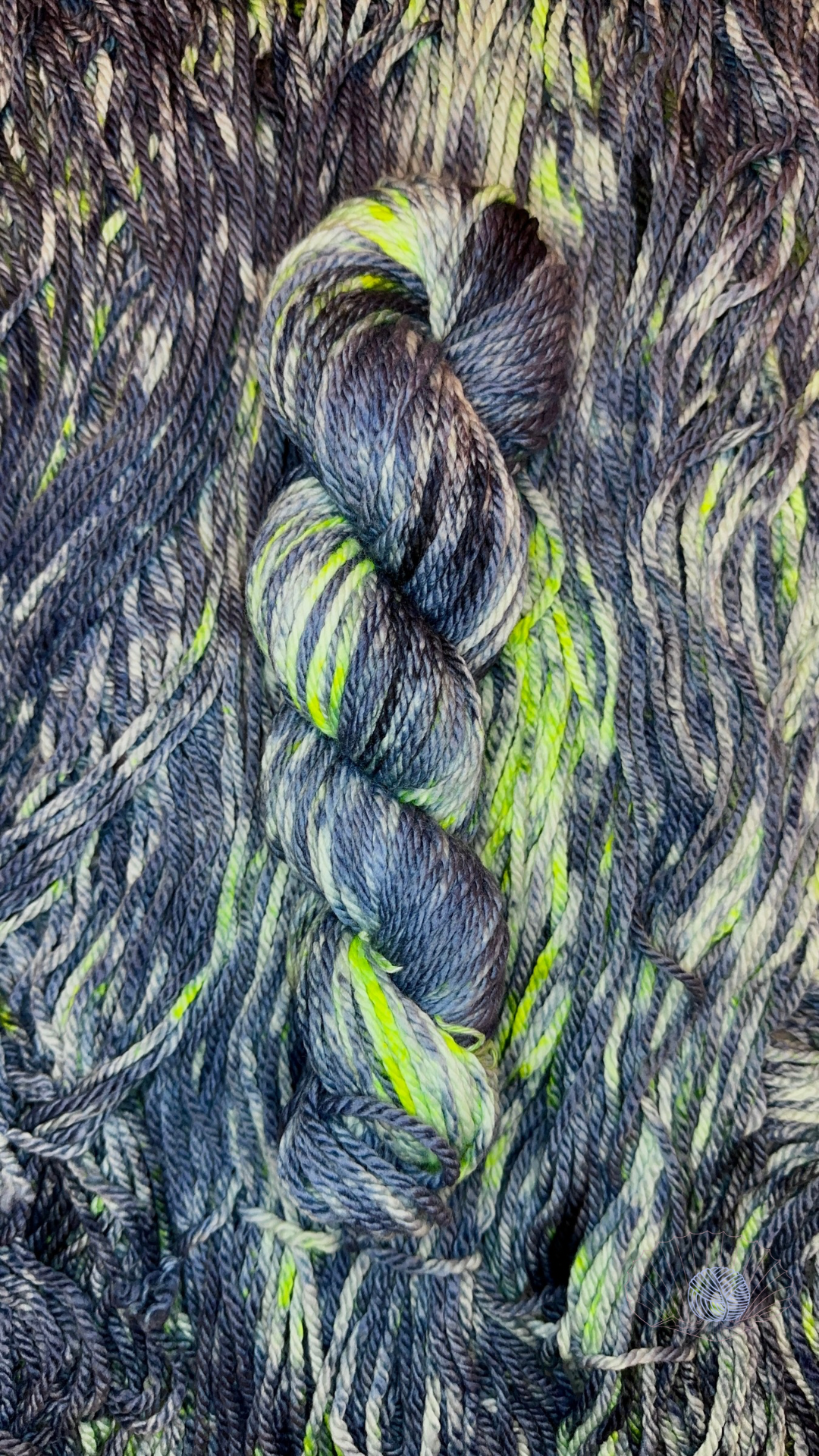 A high contrast mottled yarn of navy slate grey against pale grey, with bright spots of neon yellow-green. It’s shown twisted in a hank laying on a bed of unwound hanks of the same colorway.