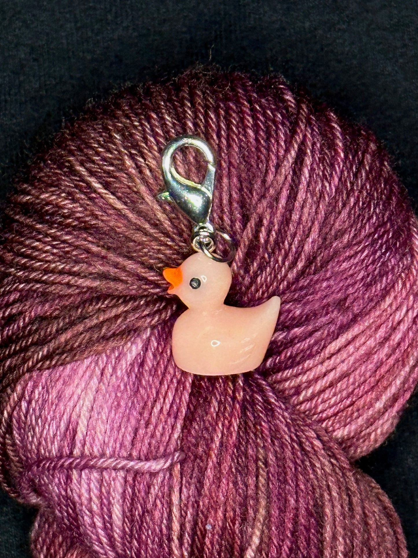 A small peachy pink rubber-duck-shaped solid plastic stitch marker with silver lobster clasp resting on a pink and burgundy yarn