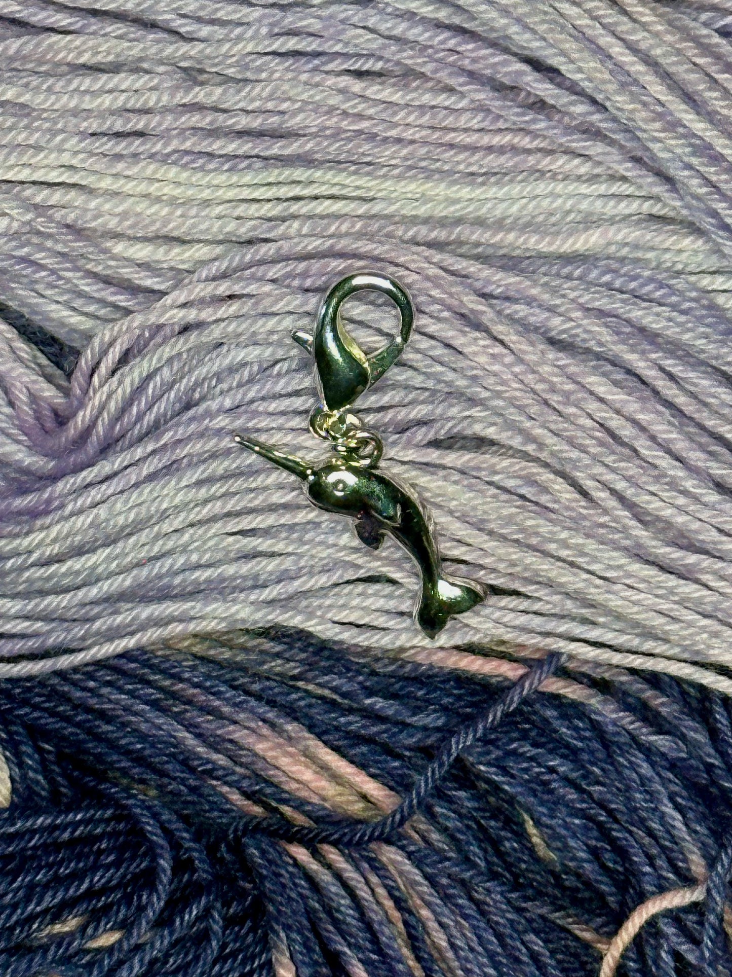 A small silver metal narwhal stitch marker with a silver lobster clasp sitting on pale lavender and navy yarn
