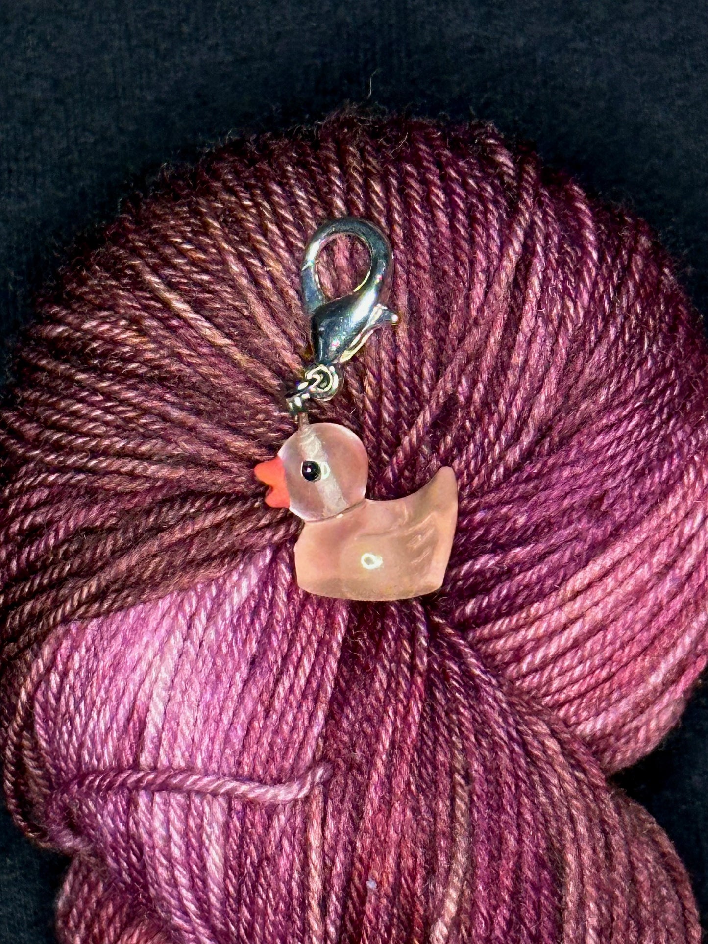 A small peachy pink rubber-duck-shaped transparent plastic stitch marker with silver lobster clasp resting on a pink and burgundy yarn