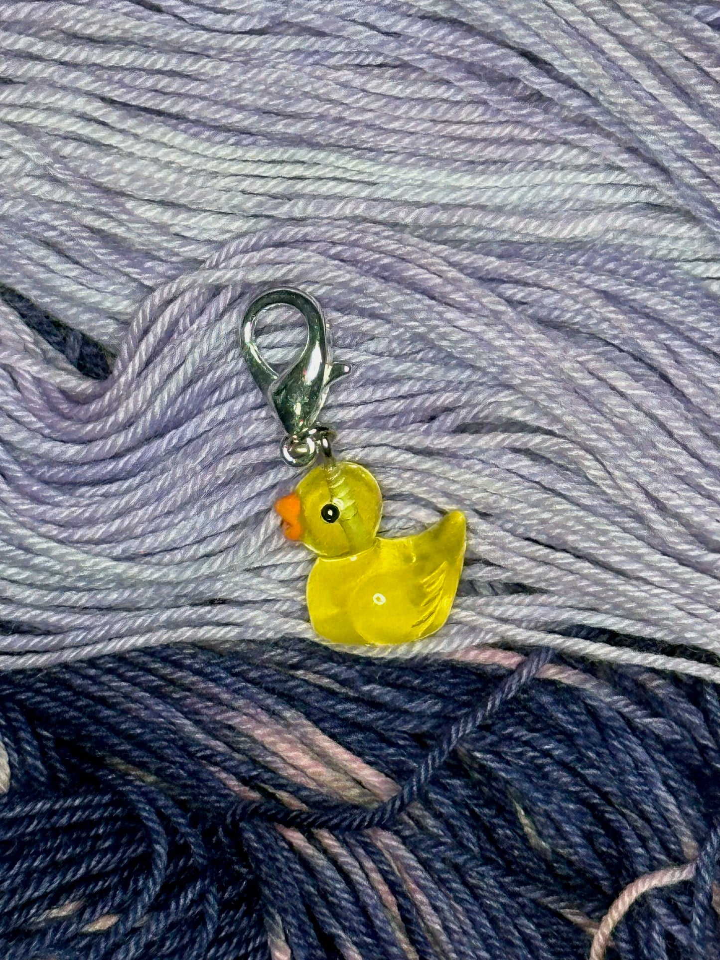 A small yellow rubber-duck-shaped transparent plastic stitch marker with a silver lobster clasp sitting on pale lavender and navy yarn