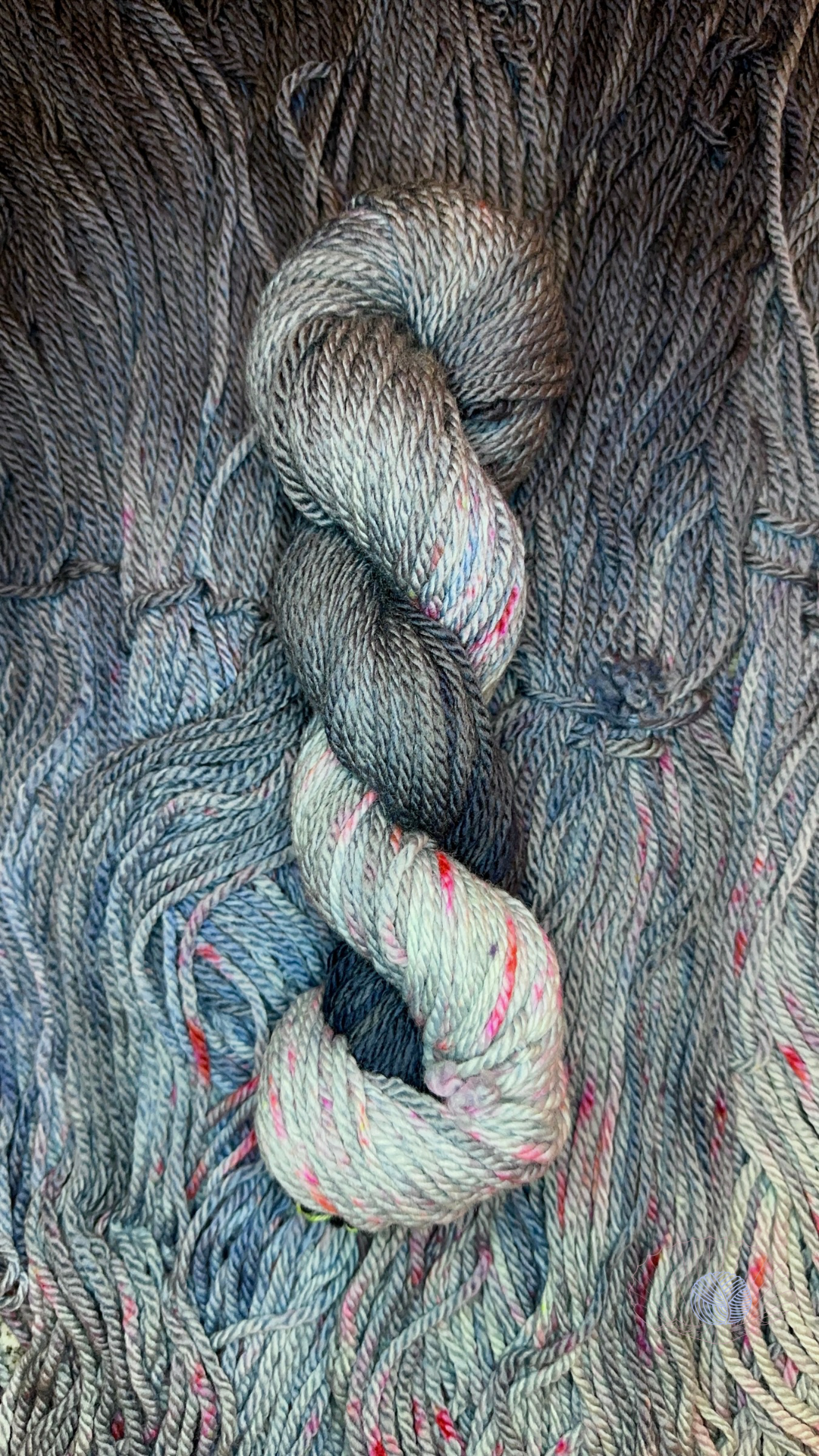 A gradient yarn colorway going from dark grey-black to pale grey with blue undertones, with bright red speckles in the pale grey section. It’s shown twisted into a hank laying atop a bed of unwound hanks of the same colorway.