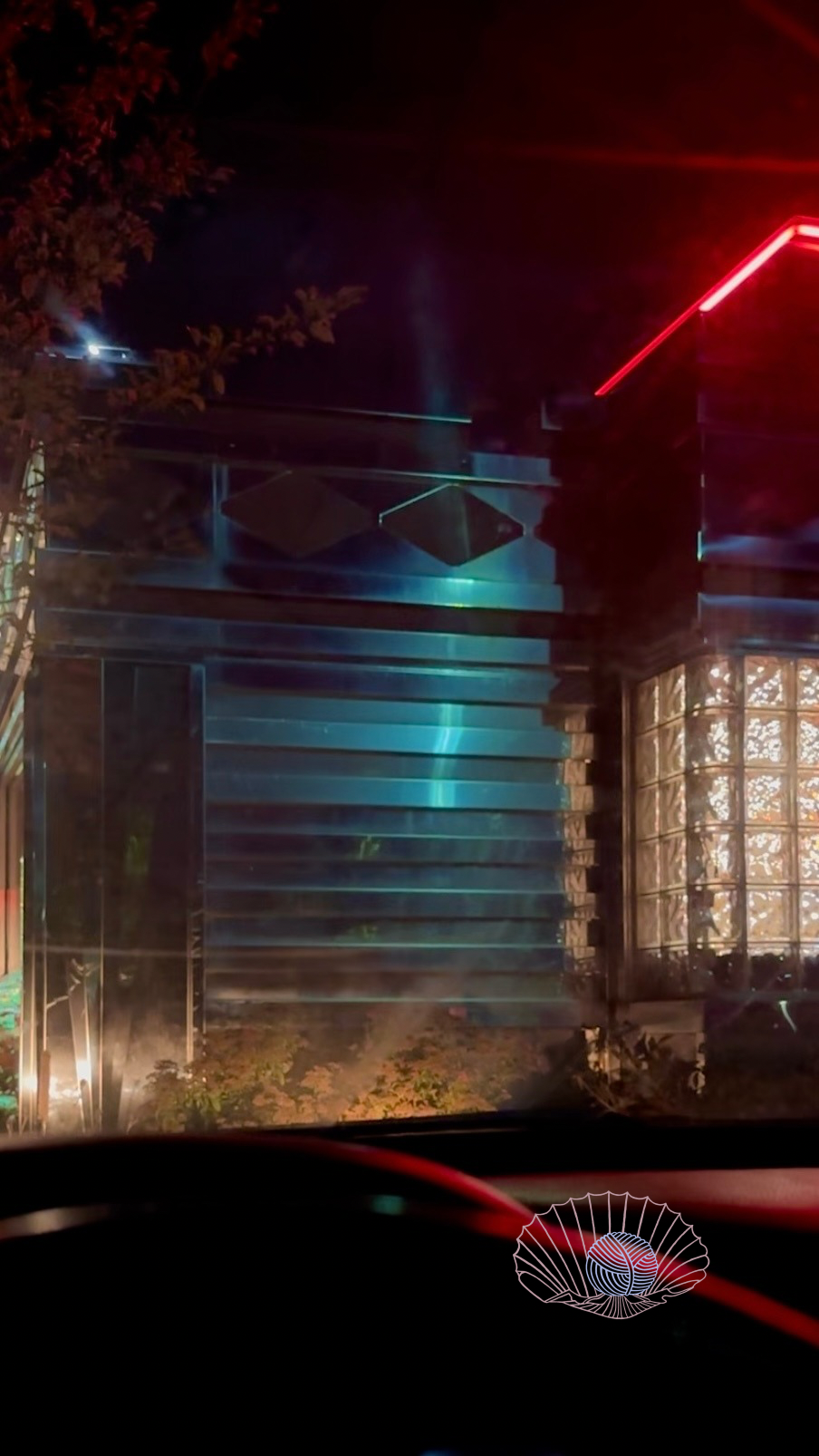 View from inside a car with a red glow on the dashboard showing the metal side of a restaurant and its neon lights.