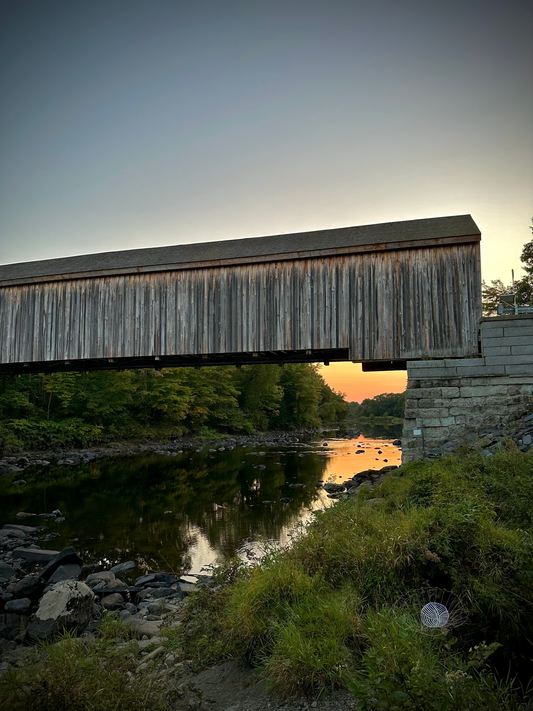 Greying wooden covered bridge over low rocky water as seen from the riverbank