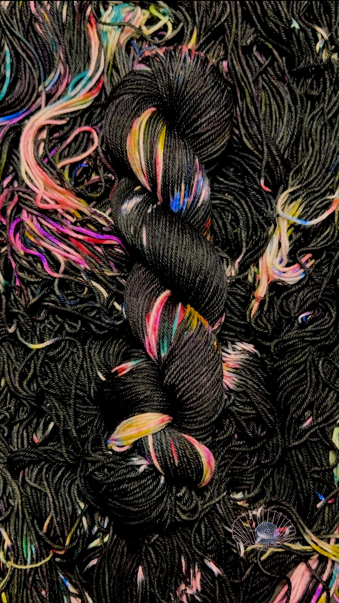 A variegated hank of yarn consisting of large black sections and randomized smaller sections of rainbow colors sitting atop a pile of untwisted hanks of the same kind.