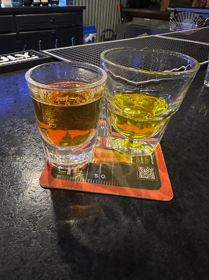 Two shot glasses sitting on a foam coaster on a black bar top. Left glass filled with whiskey and right glass filled with pickle juice.