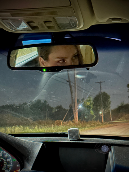 A woman’s eyes reflected in a rearview mirror looking off to the side and a road with streetlights, trees, and grassy vegetation seen through the windshield