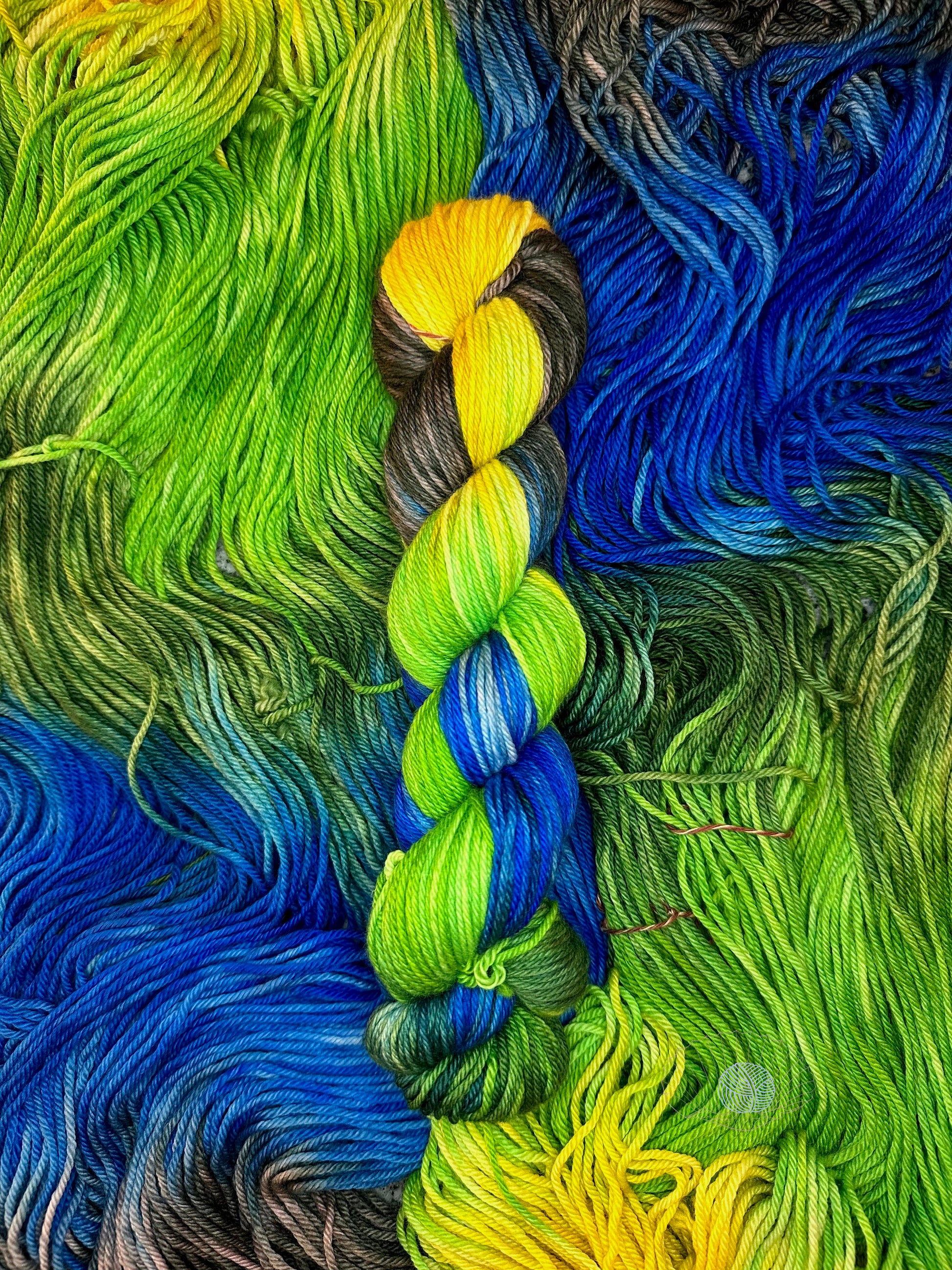 A variegated split dye worsted yarn showing opposite ends of warm yellow and dark grey asphalt at the top fading into bright green and ultramarine blue twisted together.