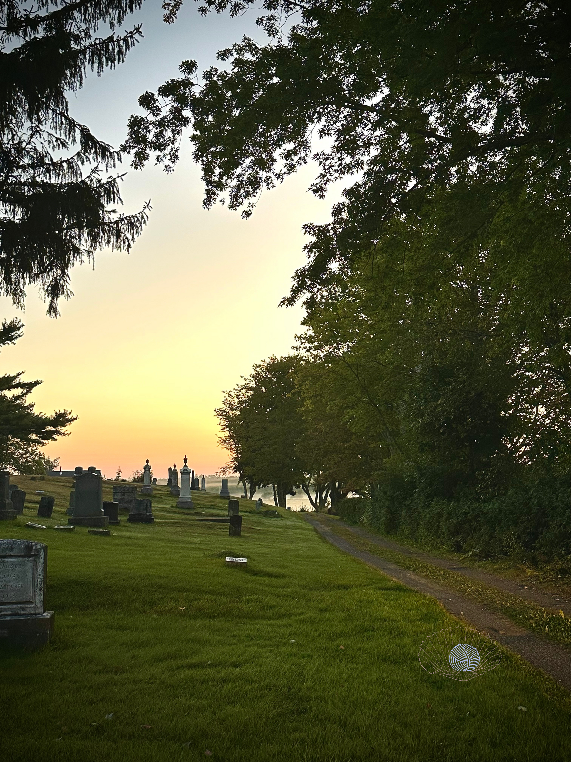 A grassy cemetery hillside with various shaped headstones with a dirt road to the right and a foggy morning field in the distant background