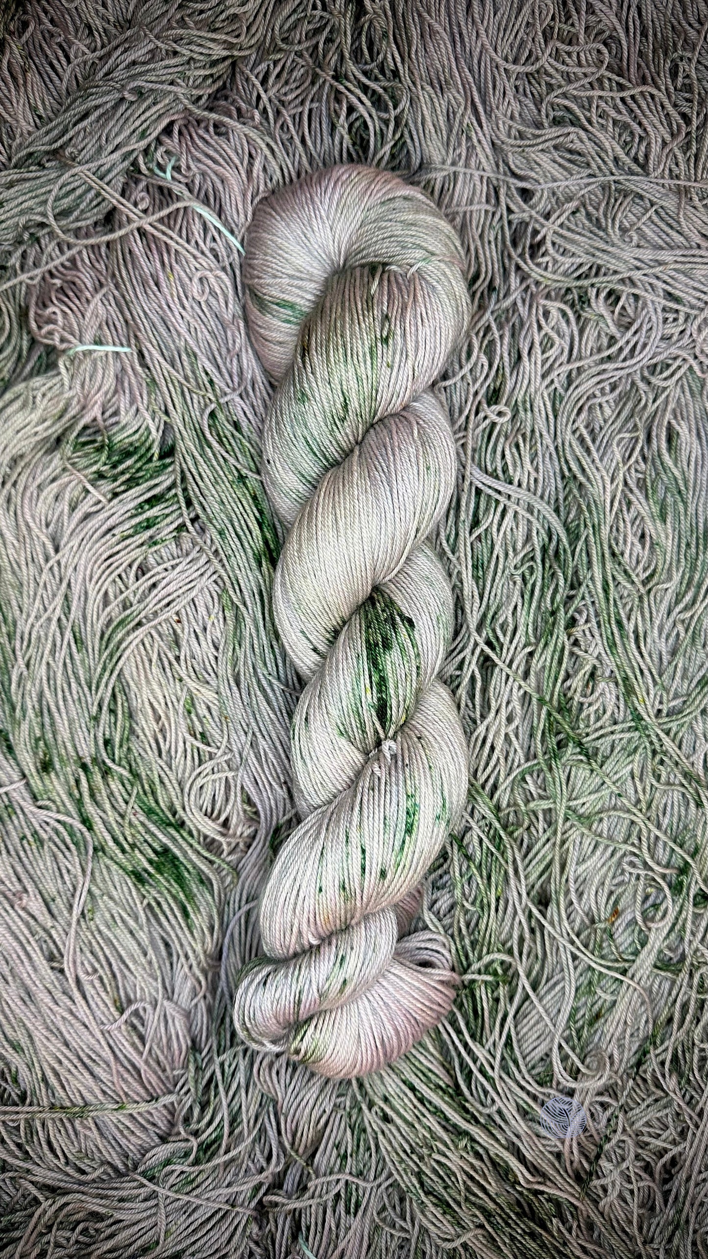 A hank of sock weight yarn in a light shade of dull grey with dark green speckles