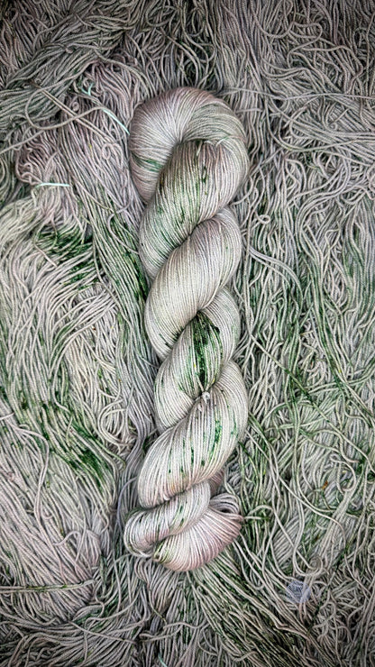 A hank of sock weight yarn in a light shade of dull grey with dark green speckles